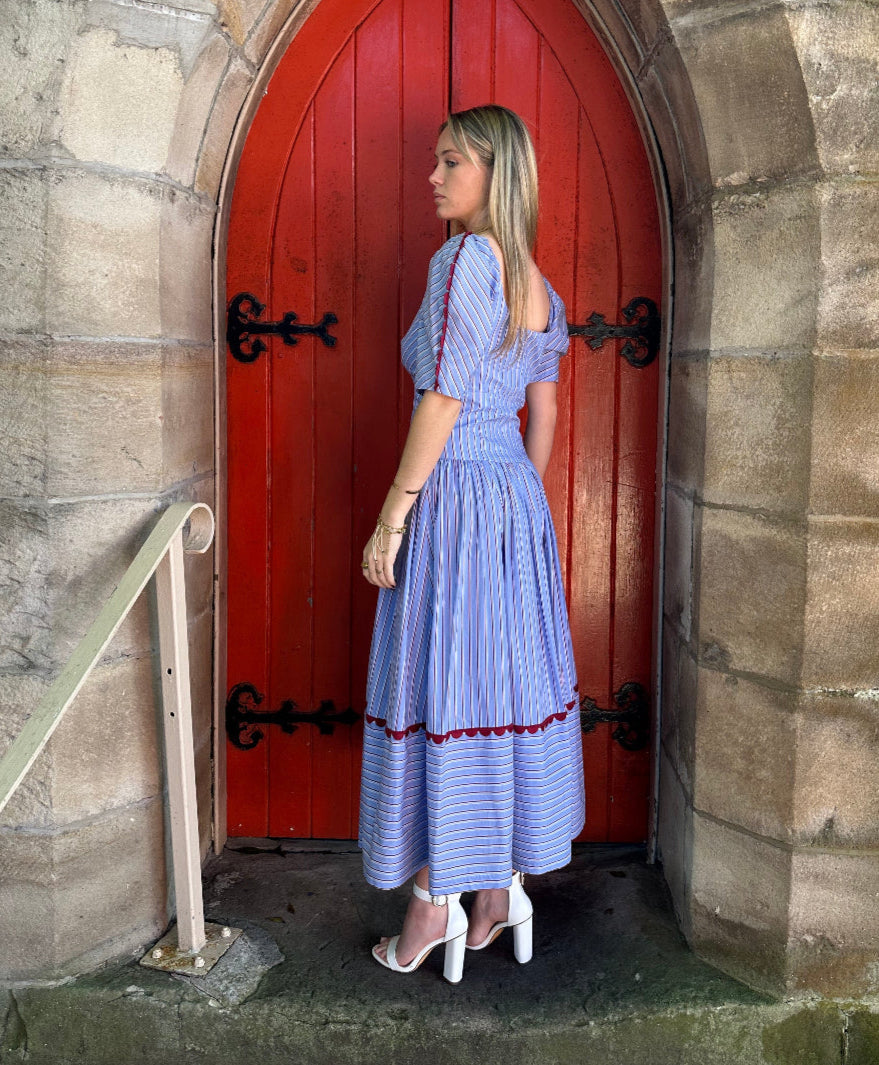 Woman in a blue dress standing inside a red door with stone walls.