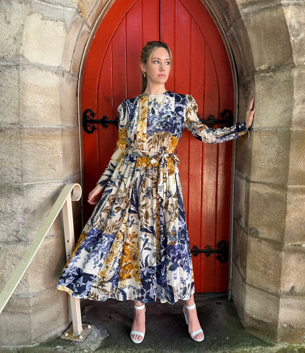 Woman in a floral dress standing in front of a red door with stone walls.