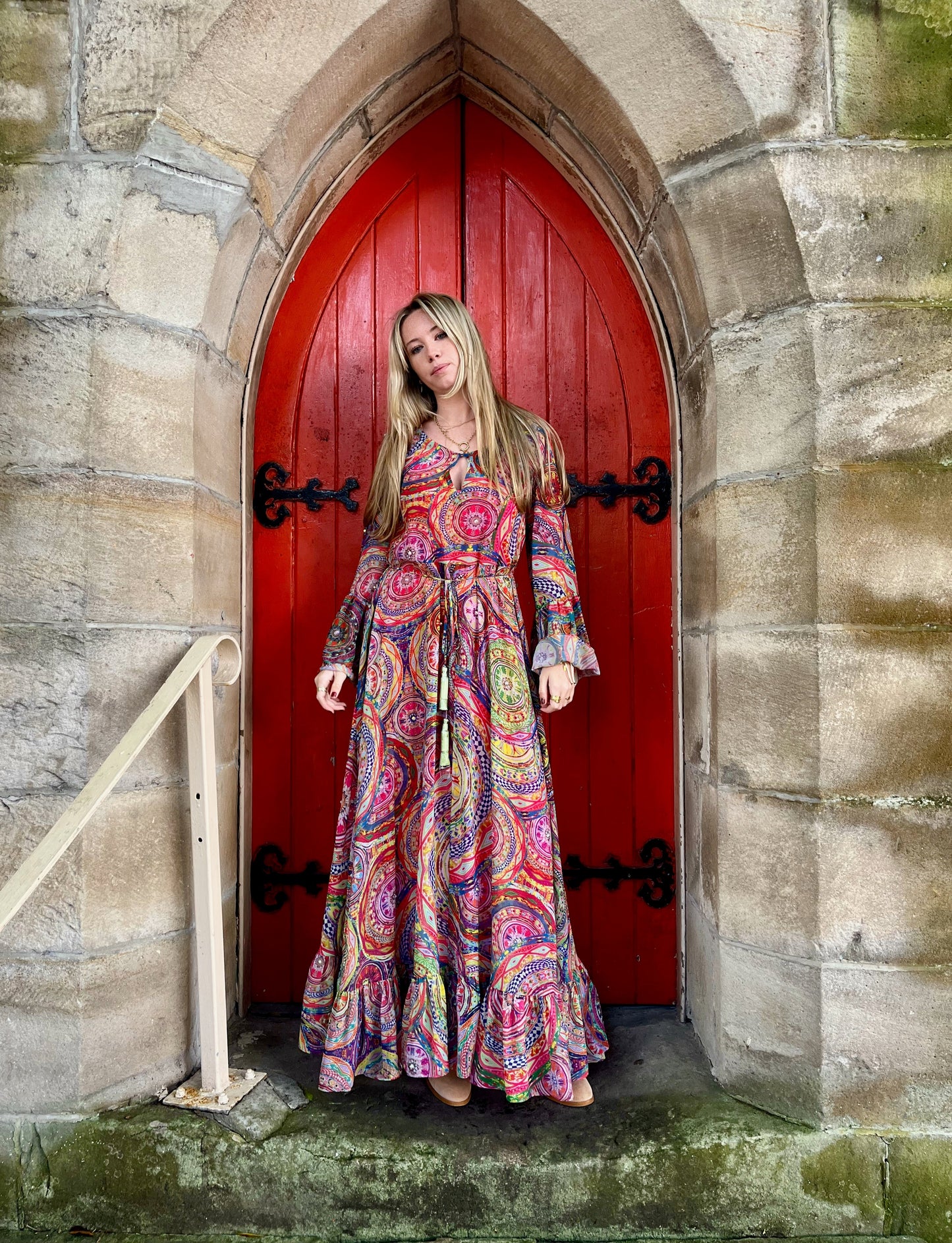 Woman in a colorful dress standing in front of a red door with stone walls.