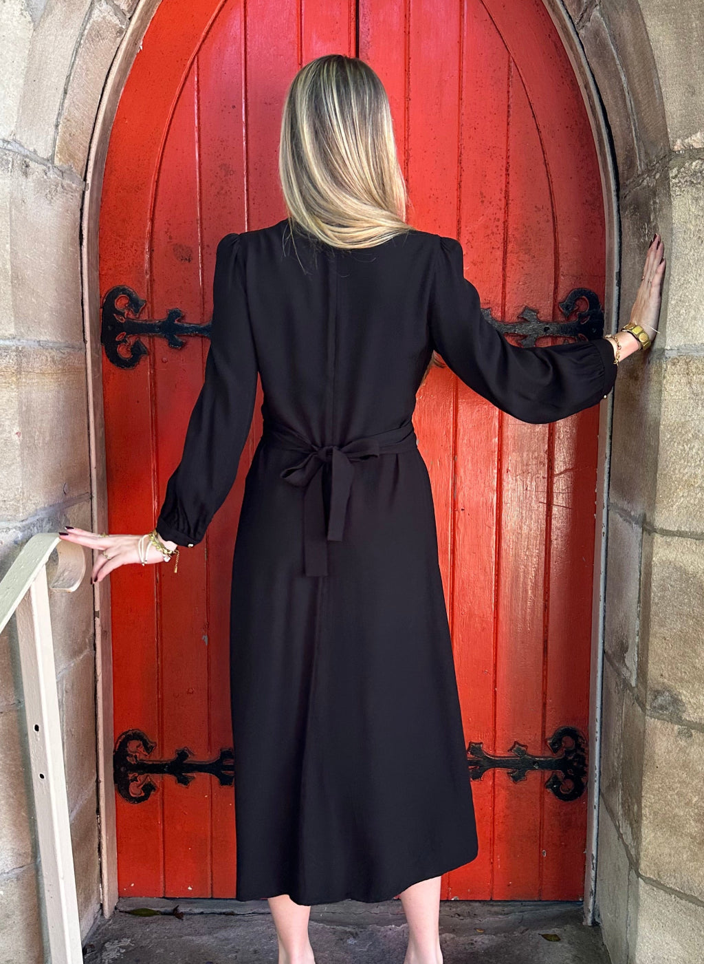Person in a black outfit standing in front of a red door on a stone wall.