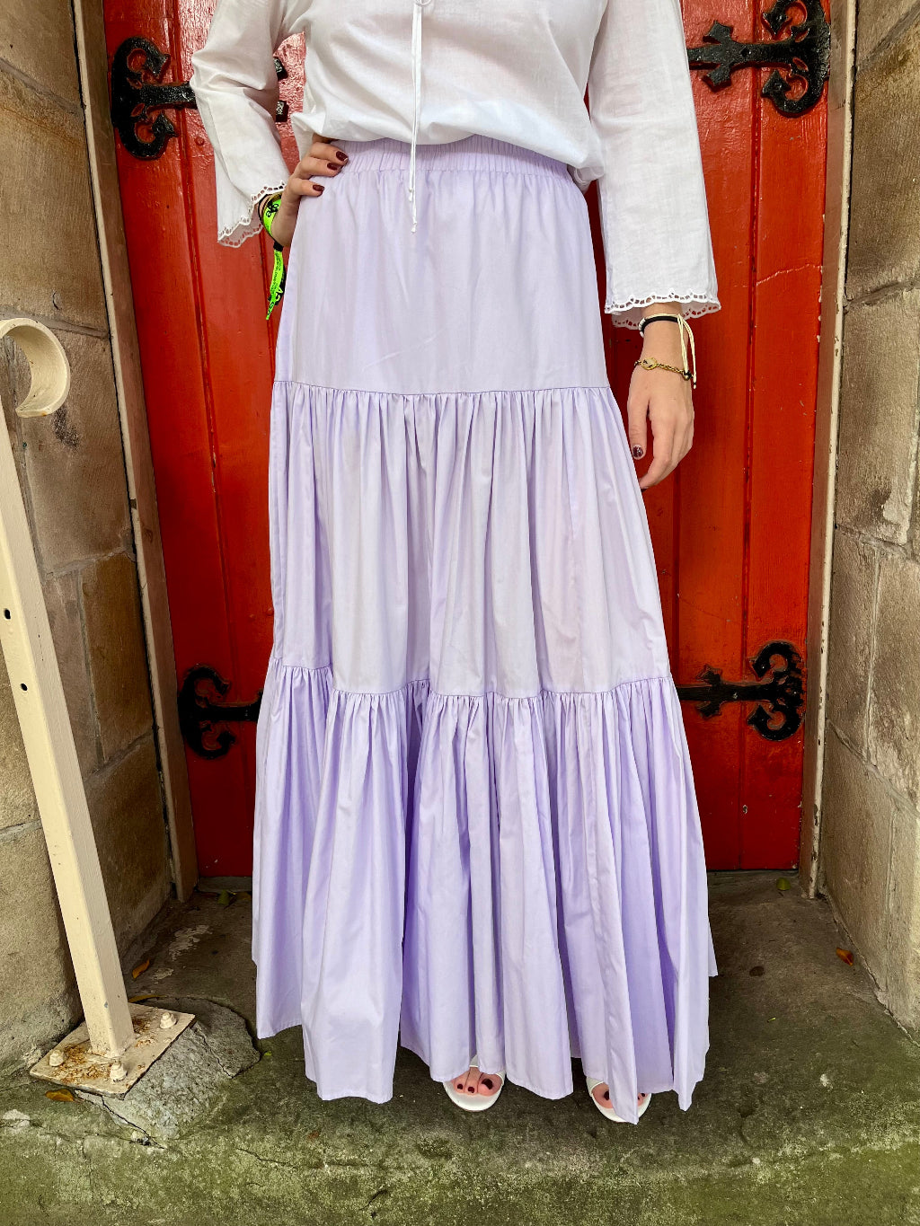 Person wearing a white top and light purple skirt standing in front of a red door.