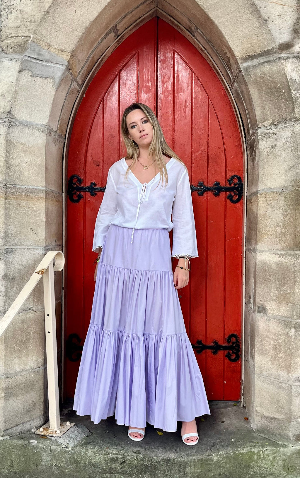 Woman in a white blouse and light purple skirt standing in front of a red door on a stone wall.