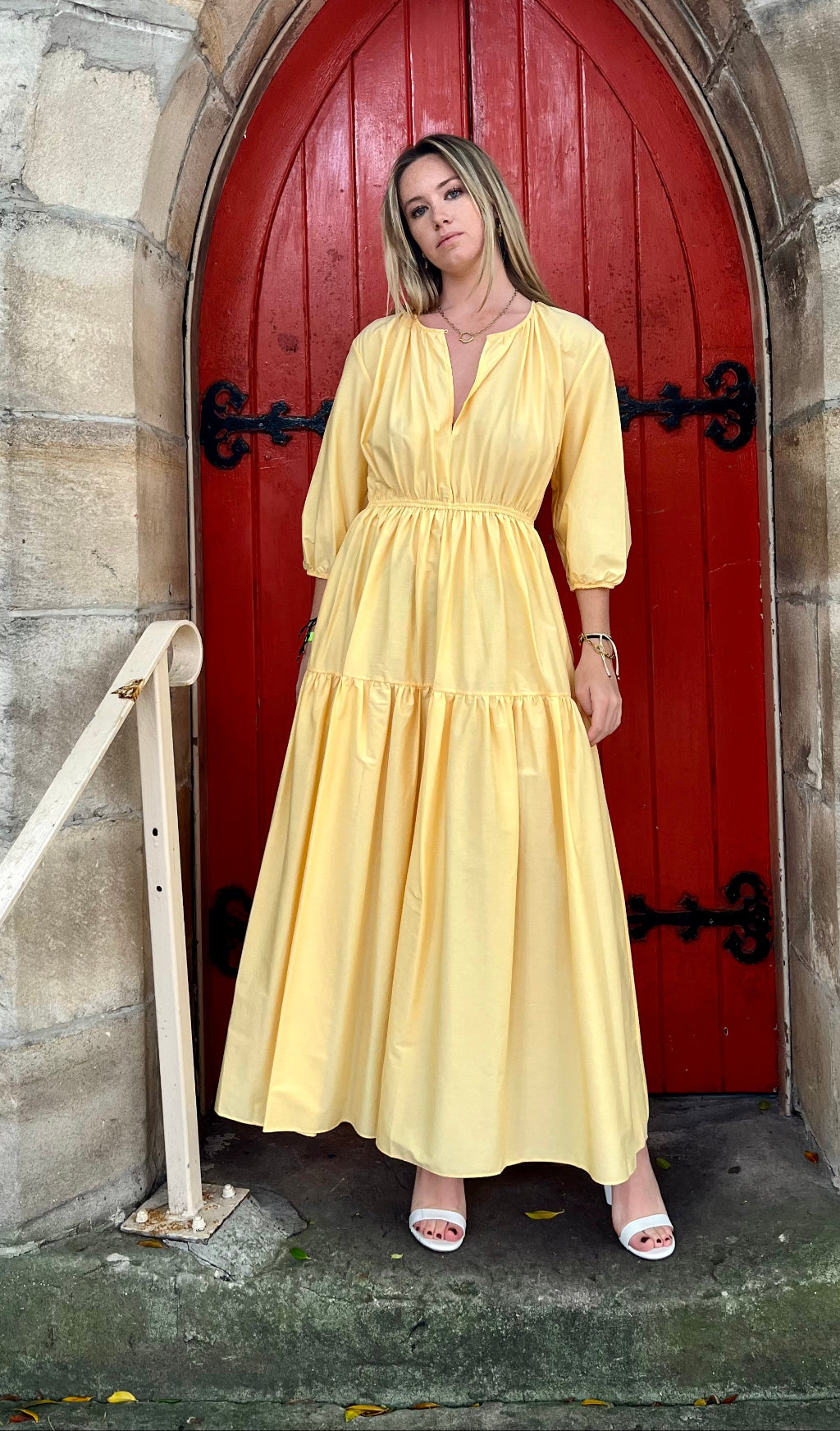 Woman in a yellow dress standing in front of a red door on a stone wall.