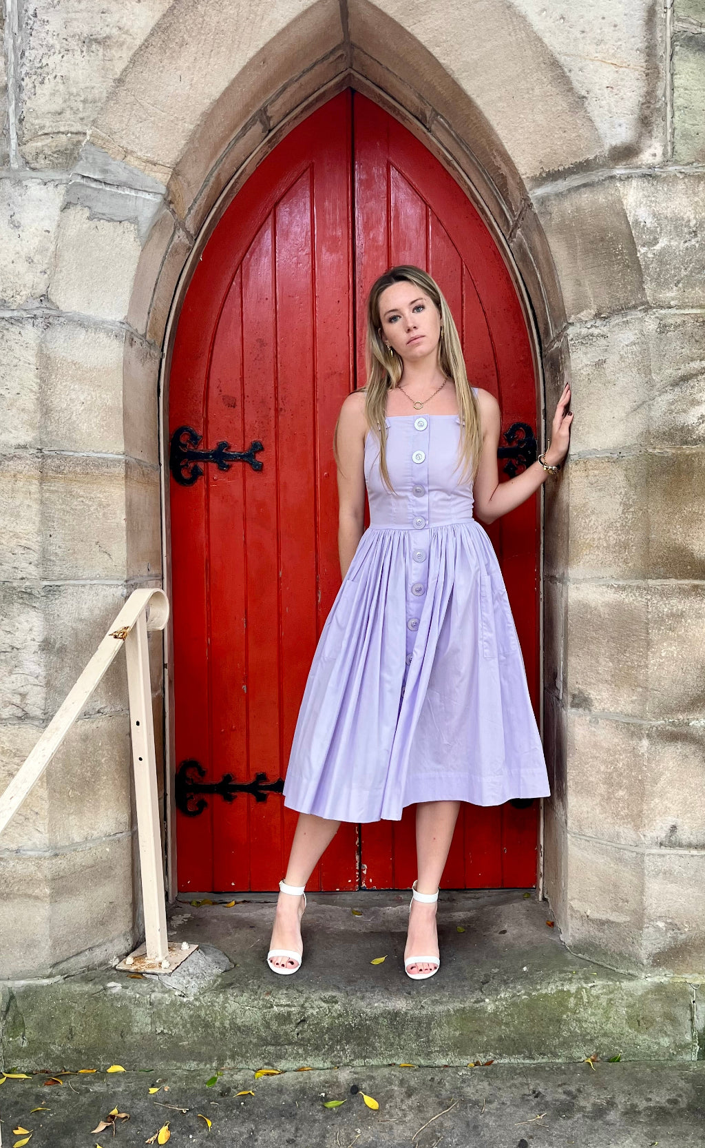 Woman in a light purple dress standing in front of a red door on stone steps.