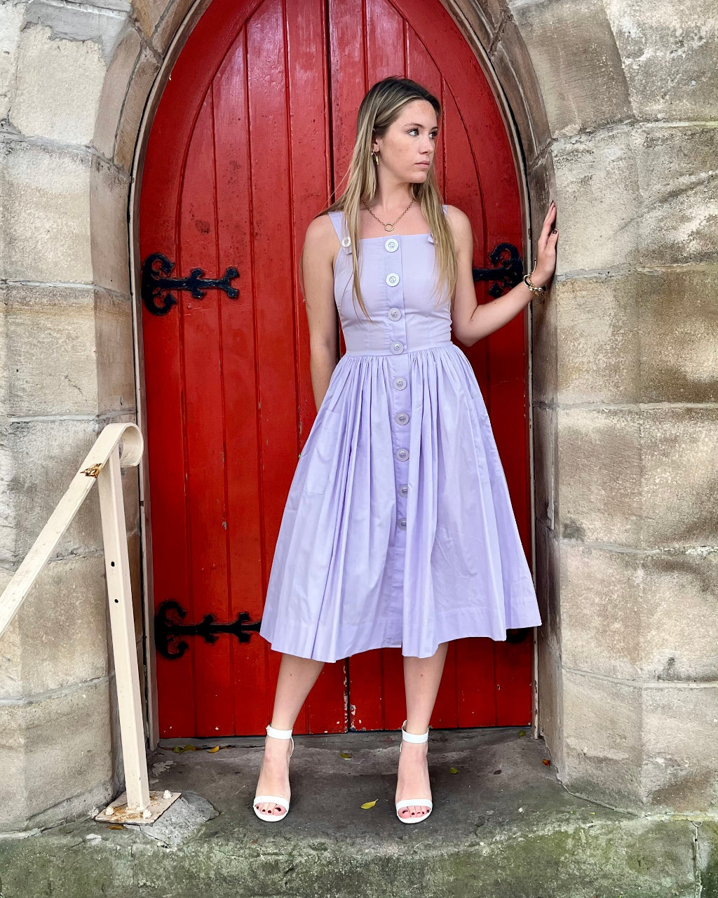 Woman in a light purple dress standing in front of a red door on stone steps.