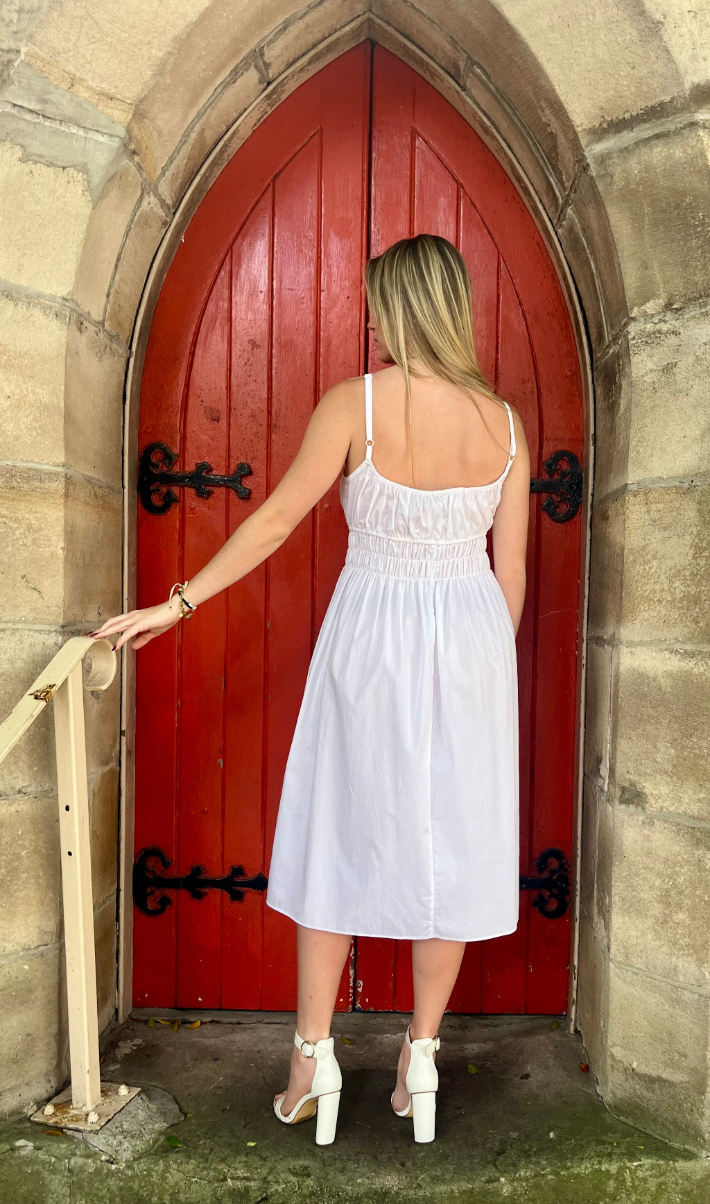 Woman in a white dress standing in front of a red door on a stone wall.