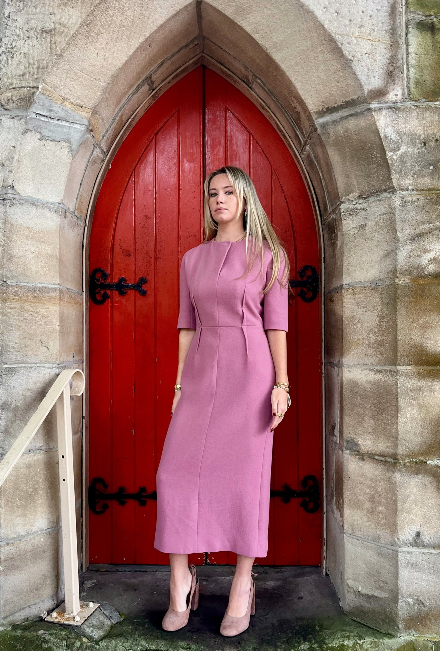 Woman in a pink dress standing in front of a red door on a stone wall.