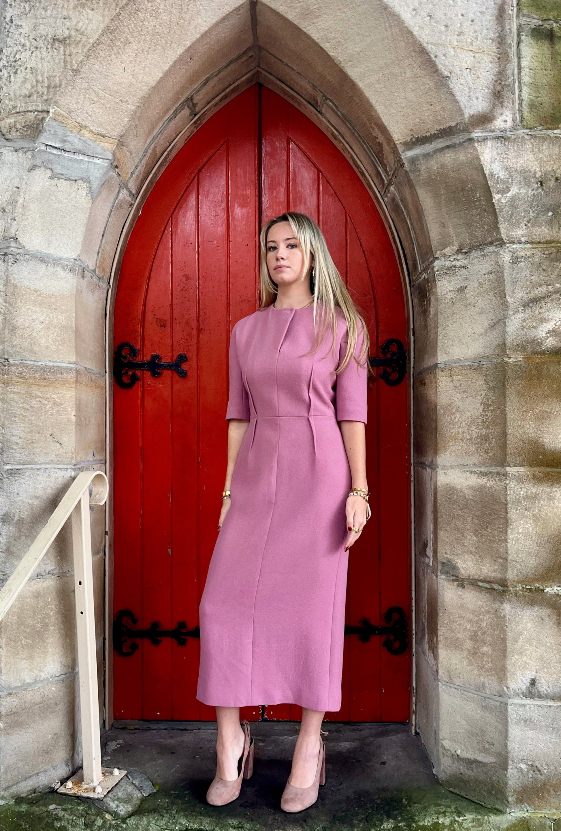 Woman in a pink dress standing in front of a red door on a stone wall.