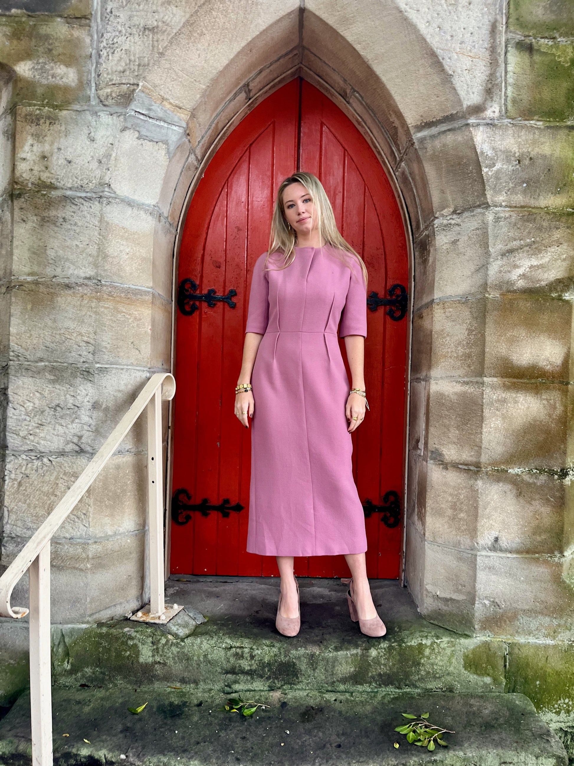 Woman in a pink dress standing in front of a red door on a stone wall.