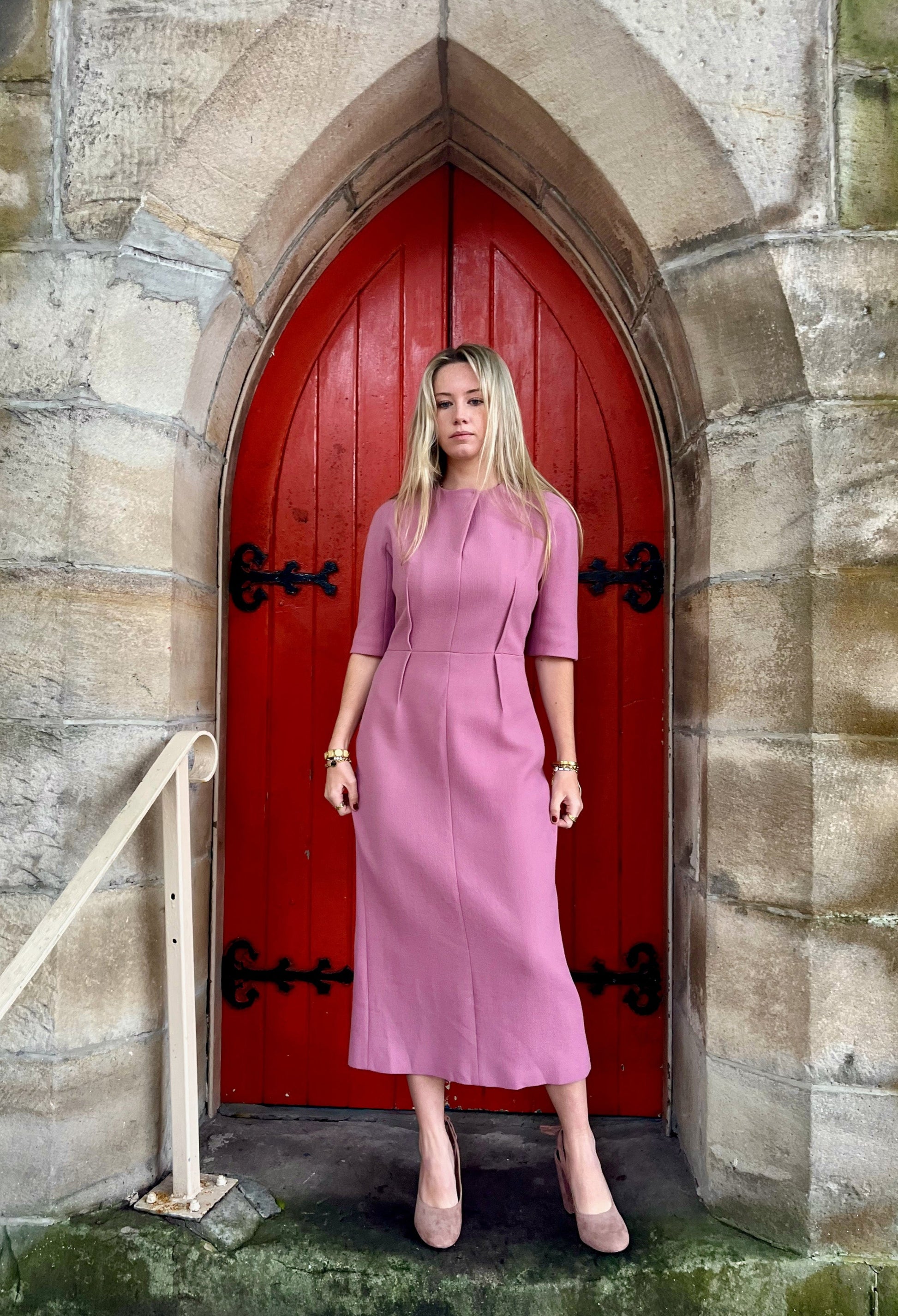 Woman in a pink  Emilia Wickstead dress standing in front of a red door on a stone wall.
