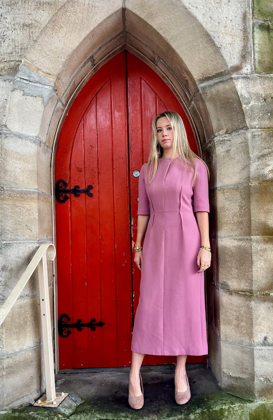 Woman in a pink  Emilia Wickstead dress standing in front of a red door on a stone wall.