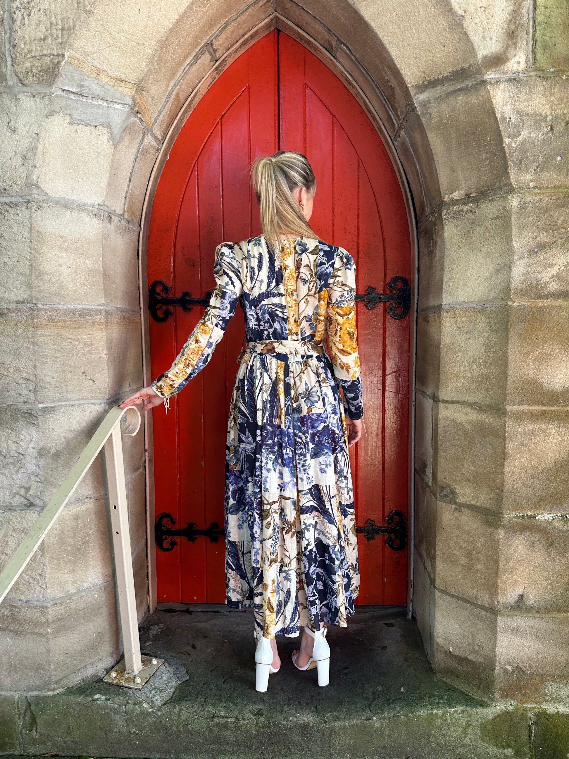 Person wearing a patterned dress standing in front of a red door with stone walls.
